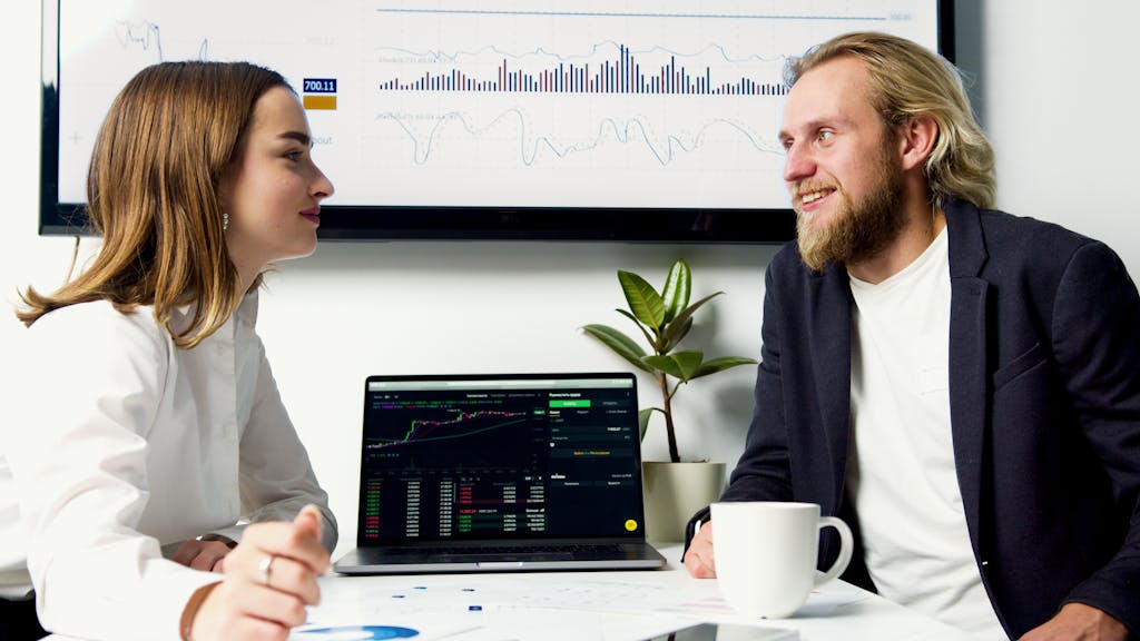 A business meeting with two professionals discussing data on laptops with charts displayed. financial planning