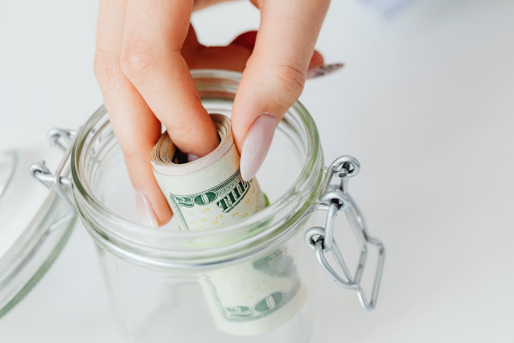 A close-up of a hand placing rolled dollars into a glass jar, symbolizing savings. emergency funds