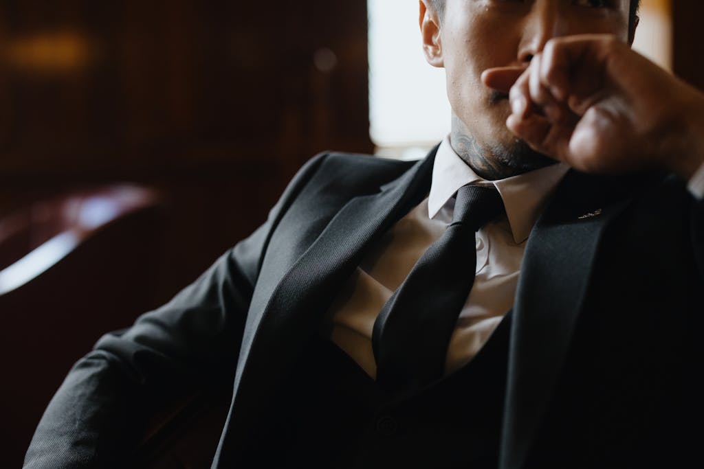 A man in a black suit sitting indoors with a thoughtful expression, hand on face. ex has moved on. learn how to tie a tie like his