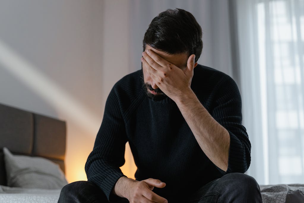 A man in distress sits on a bed indoors, covering his face with his hand. men's mental health awareness