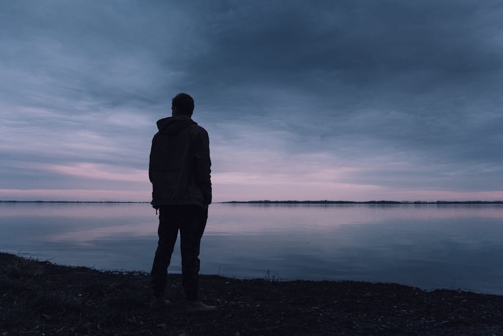 A solitary silhouette of a man in a jacket gazing at a lake during a peaceful sunset, creating a serene atmosphere. men's mental health awareness