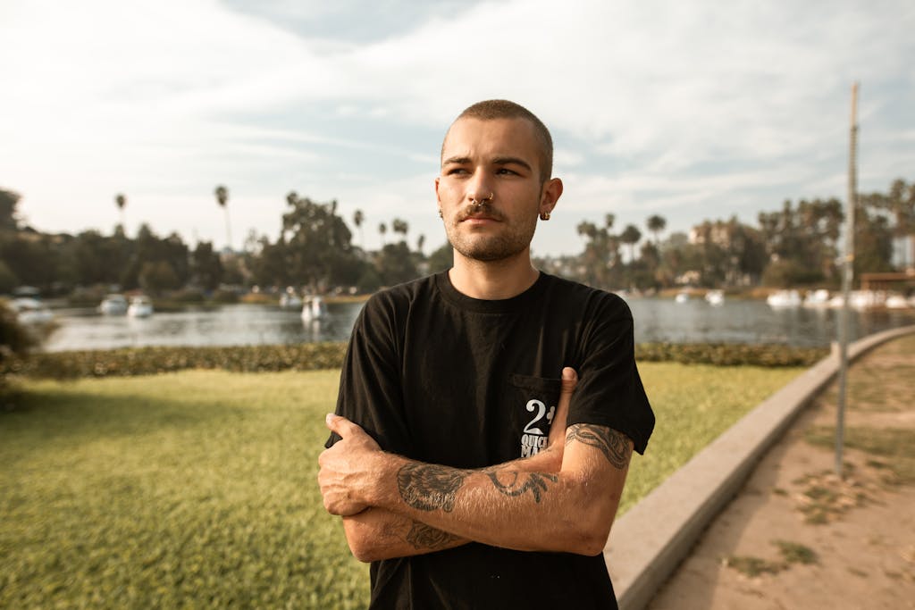 A tattooed young man stands confidently by the lake in a casual setting, exuding style and attitude. girls like bad boys