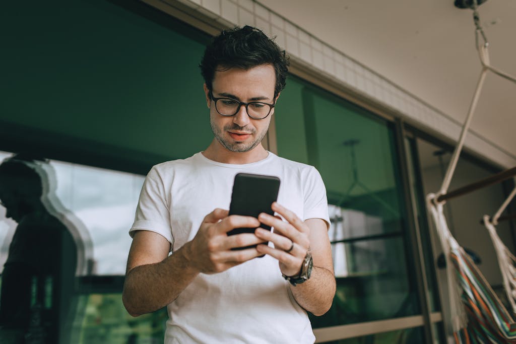 Young man standing on a balcony, using a smartphone, reflecting in the window. social media addiction